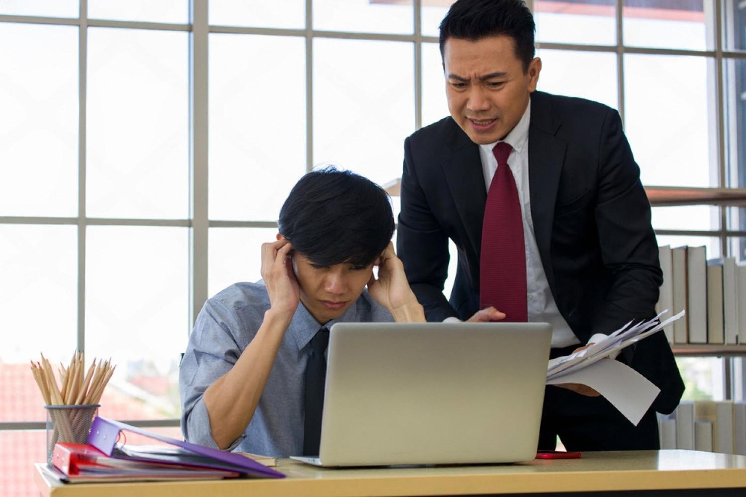 A stressed junior employee at a laptop while a manager stands over him pointing at the screen